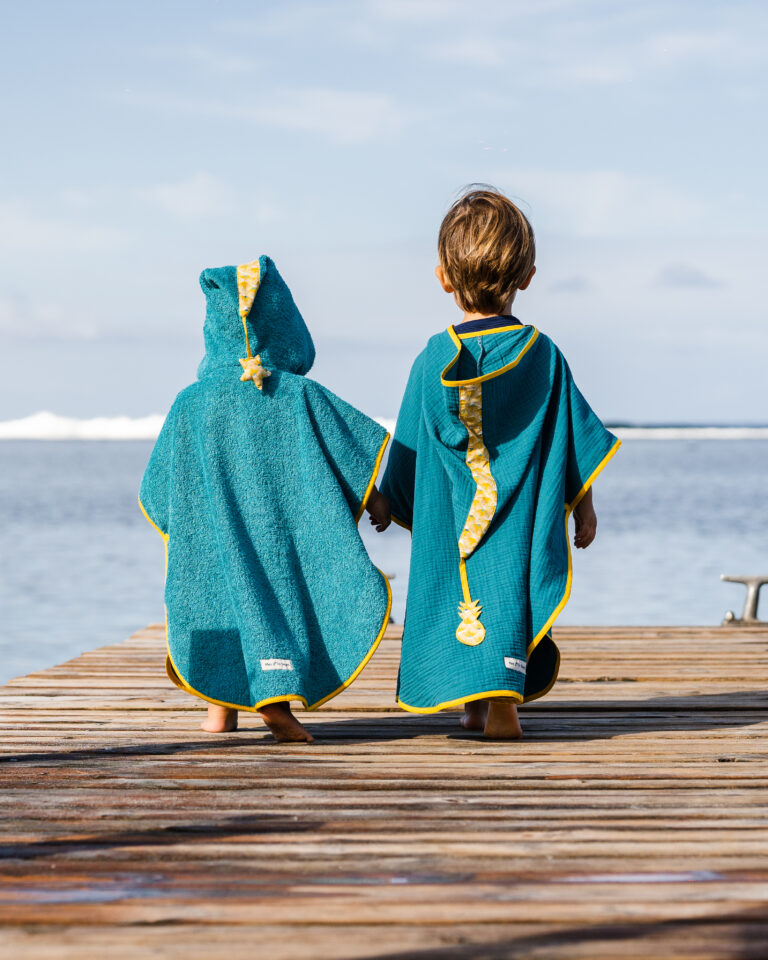 Deux enfants vus de dos, marchant sur un ponton en bois au bord de la mer, portant des ponchos de bain artisanales.