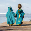 Deux enfants vus de dos, marchant sur un ponton en bois au bord de la mer, portant des ponchos de bain artisanales.