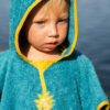 Enfant au regard sérieux portant un poncho de bain turquoise avec une broderie d'ananas jaune.