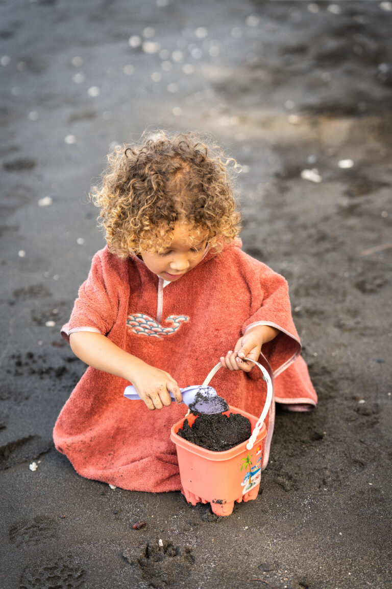 Un enfant jouant dans le sable noir avec un seau et une pelle, vêtu d'un poncho de bain rouge artisanale.