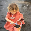 Un enfant jouant dans le sable noir avec un seau et une pelle, vêtu d'un poncho de bain rouge artisanale.