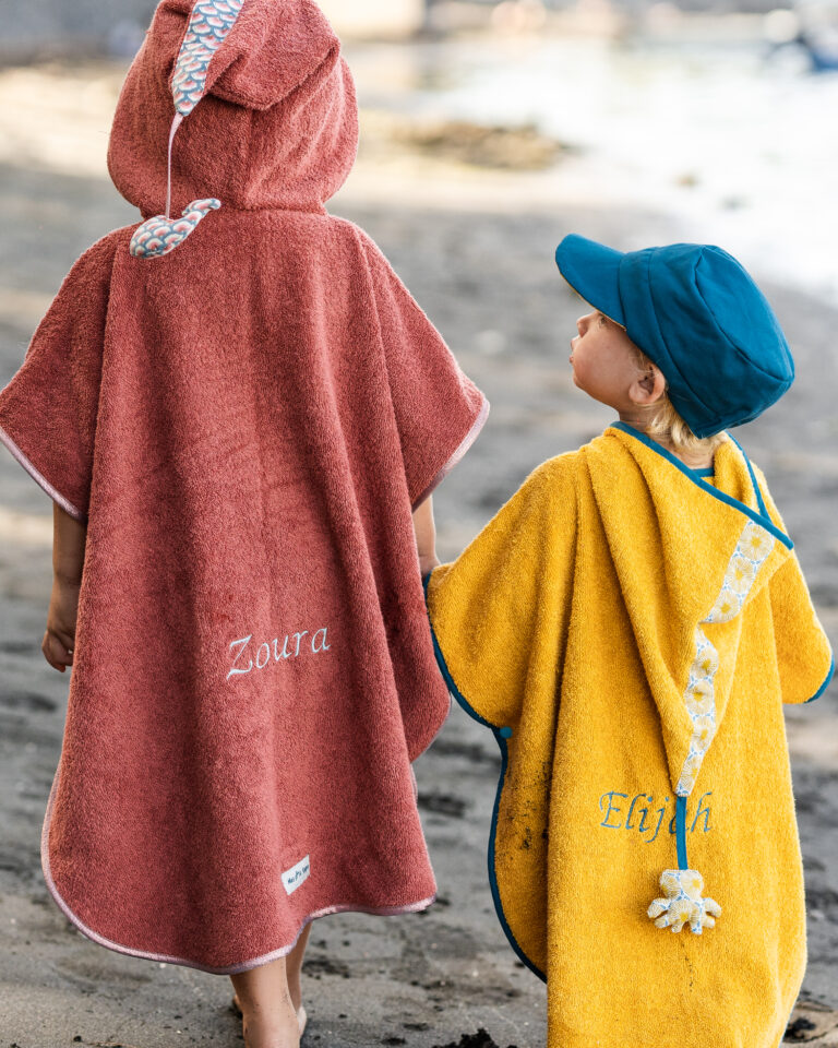 Deux enfants en poncho de bain personnalisées avec leurs noms brodés, se tenant sur une plage.