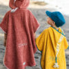 Deux enfants en poncho de bain personnalisées avec leurs noms brodés, se tenant sur une plage.