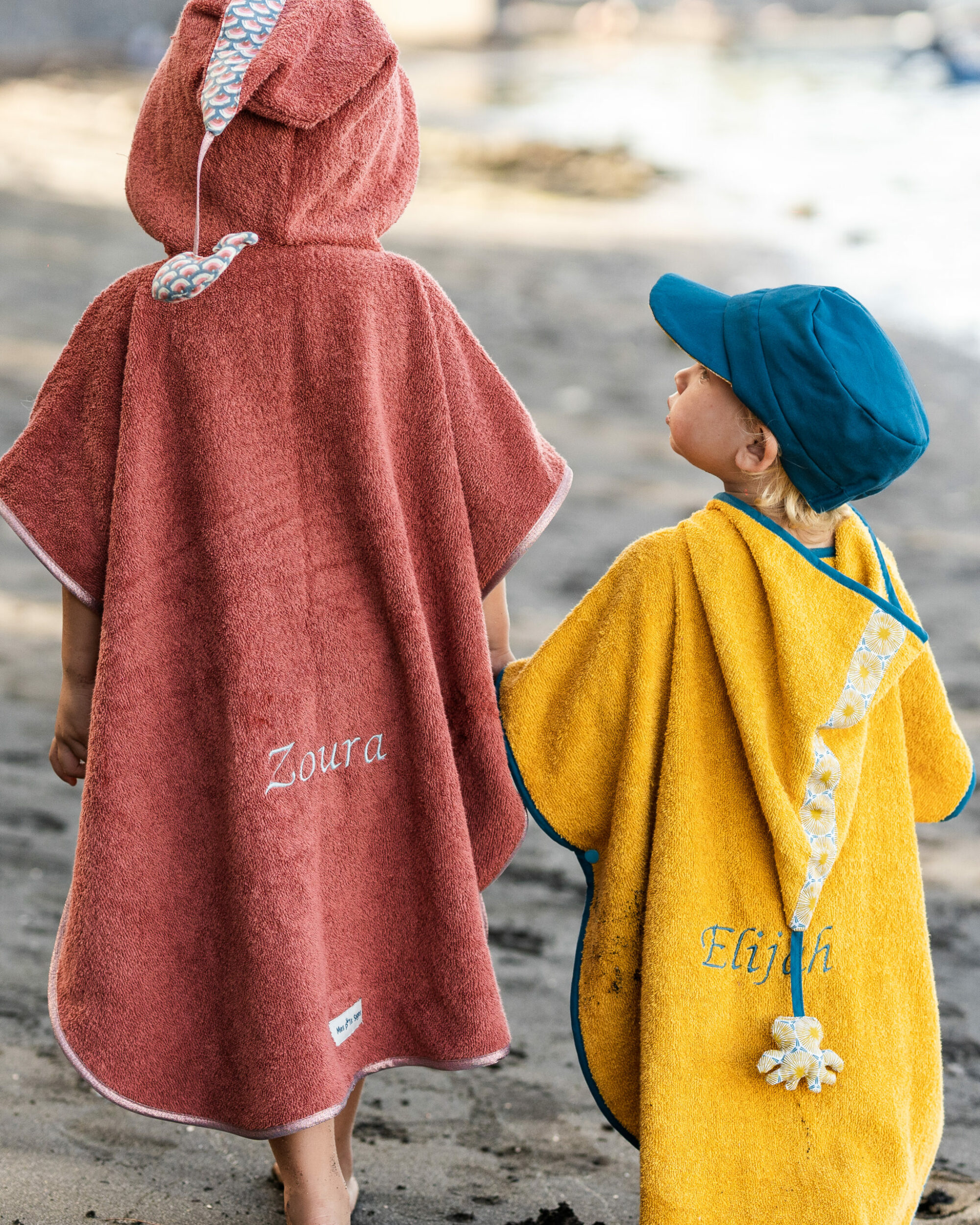 Deux enfants en poncho de bain personnalisées avec leurs noms brodés, se tenant sur une plage.