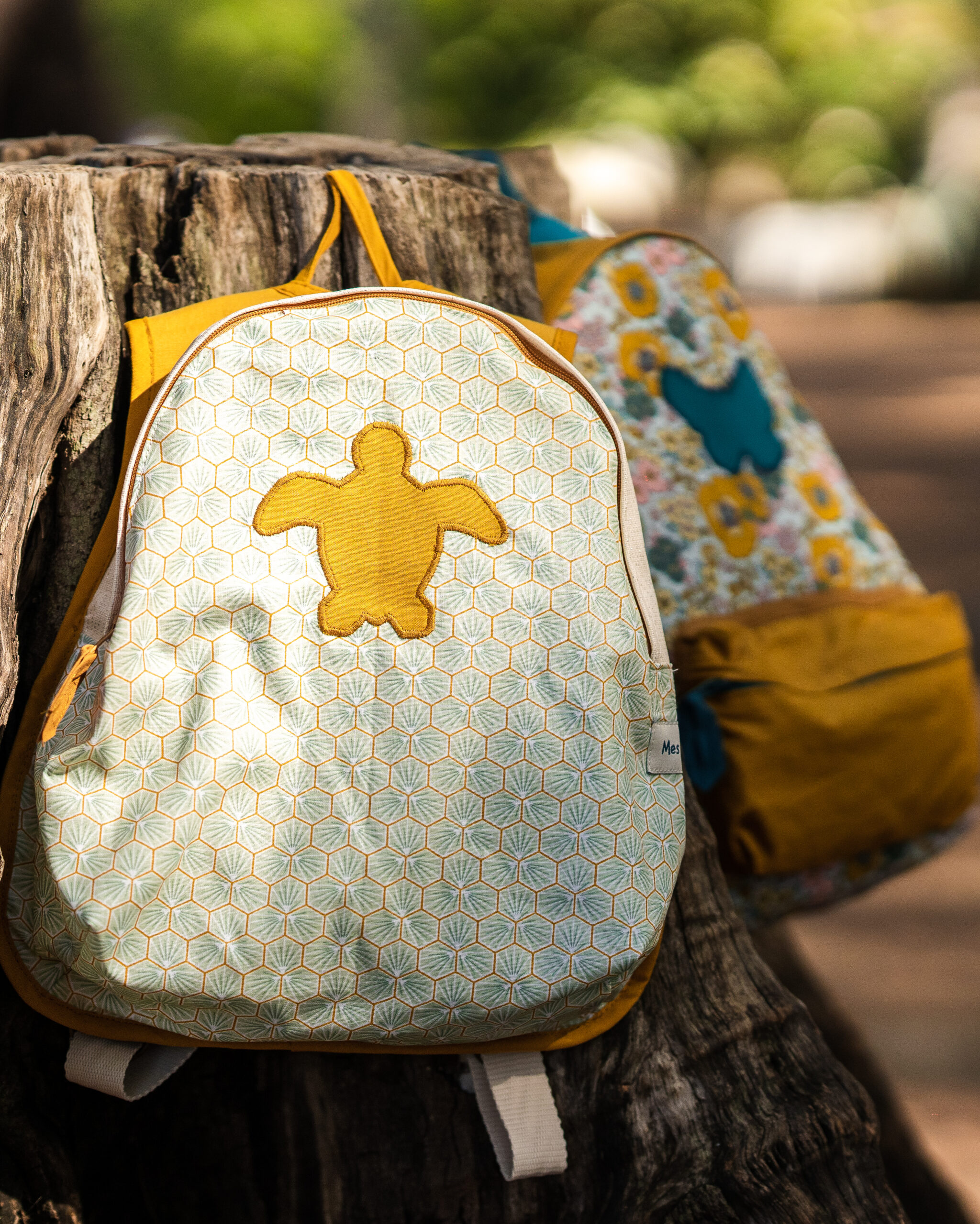 Un sac à dos enfantin avec un motif de tortue placé sur un tronc d'arbre, évoquant l'artisanat local.