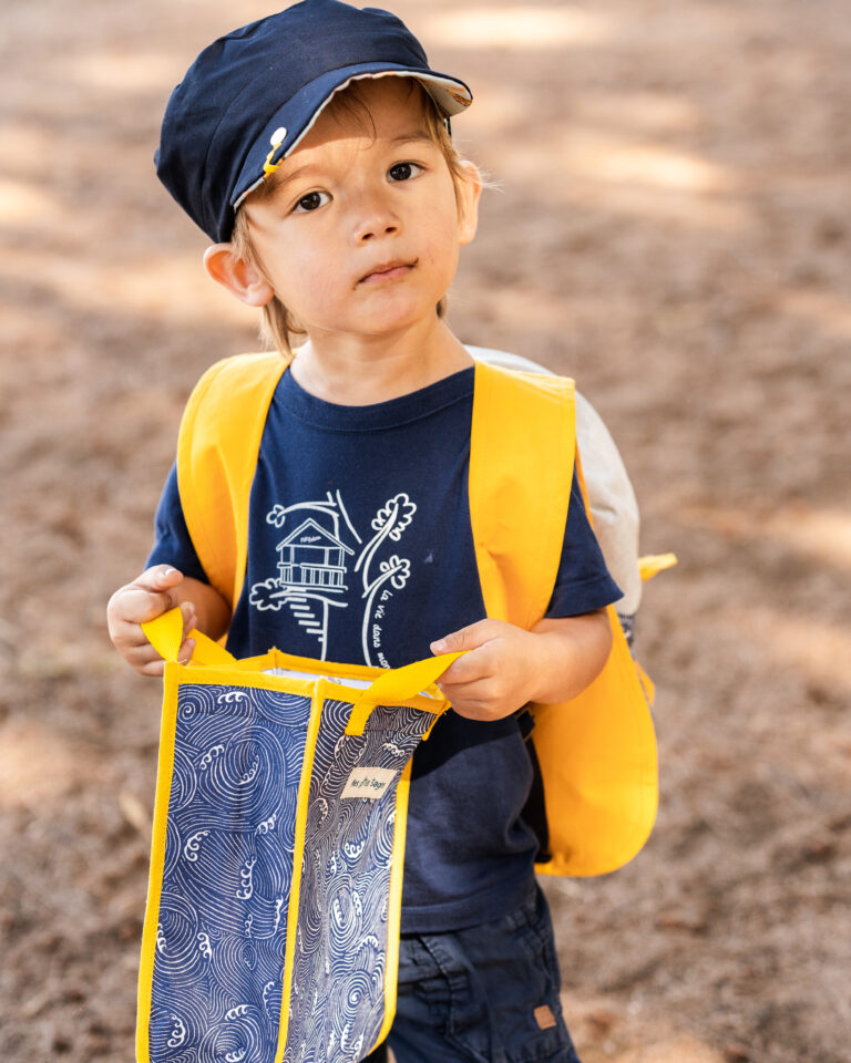Un petit garçon avec une casquette et un sac à dos jaune regardant sérieusement.