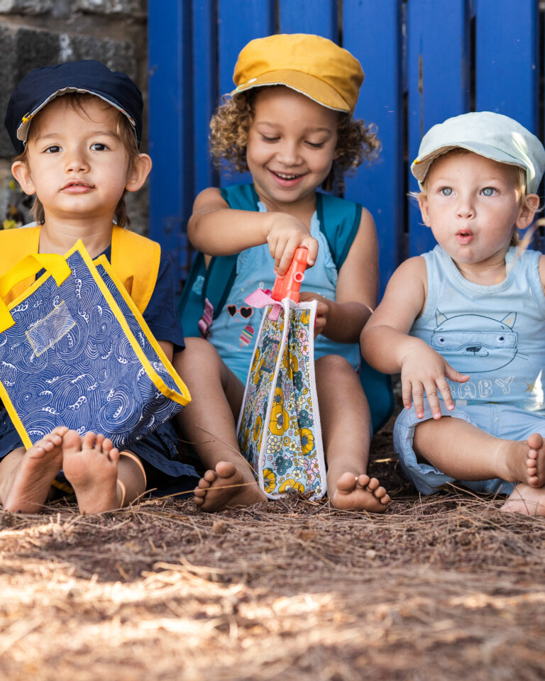 Trois enfants assis par terre, jouant et interagissant avec des sacs à dos artisanaux.