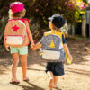 Deux enfants marchant main dans la main, avec des sacs à dos artisanaux, sur un chemin entouré de verdure.