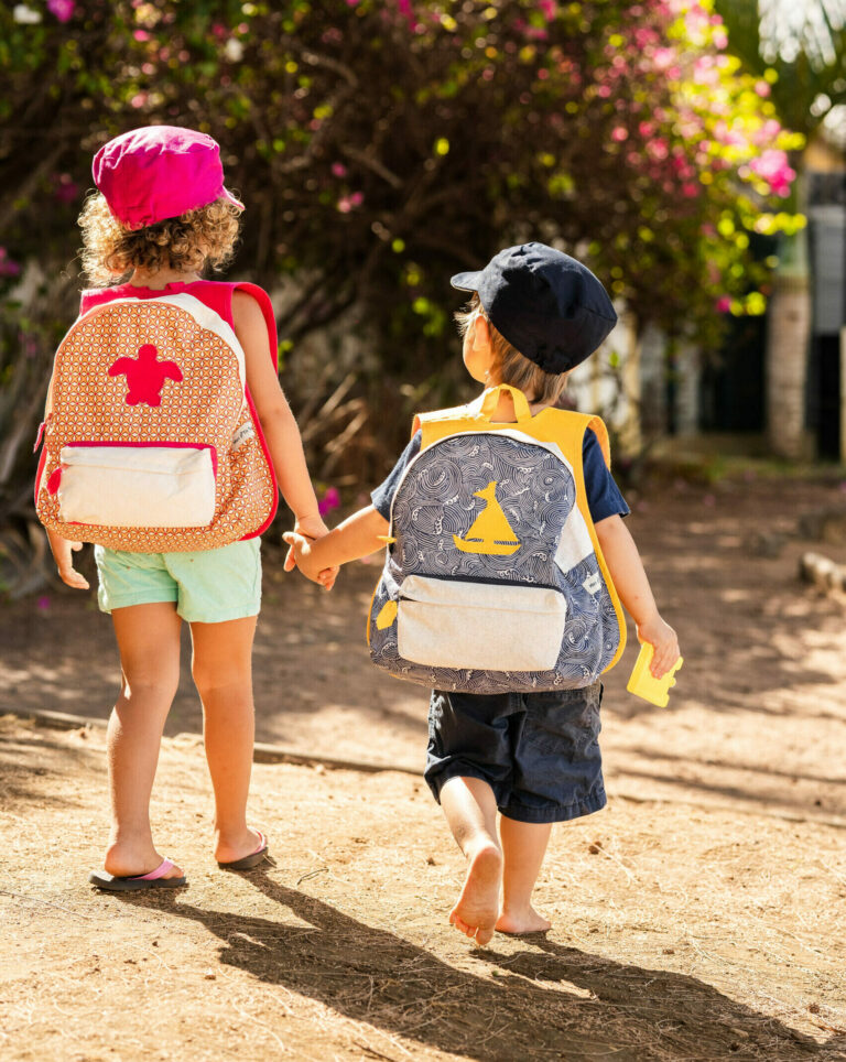 Deux enfants marchant main dans la main, avec des sacs à dos artisanaux, sur un chemin entouré de verdure.