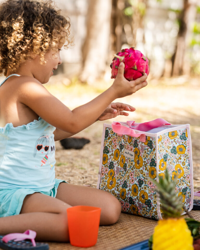 Un enfant assis dehors jouant avec un fruit tropical et un sac à motifs colorés à ses côtés.