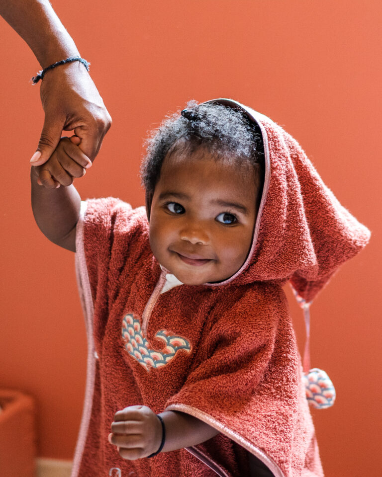 Un enfant souriant tenant la main de quelqu'un hors de l'image, vêtu d'un poncho de bain rouge avec un motif de baleine brodé.